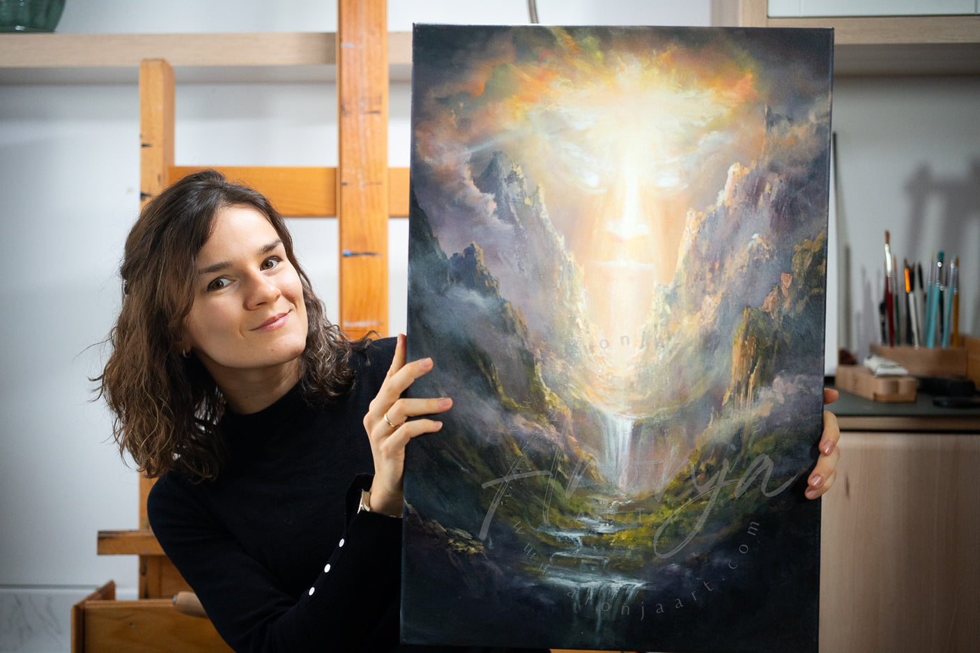 Female fantasy artist holding hand-painted SORCERY Gothic Original Painting "One True God" - 60x40cm / Acrylics featuring a mystical glowing face over a mythical landscape with waterfalls and mountains.