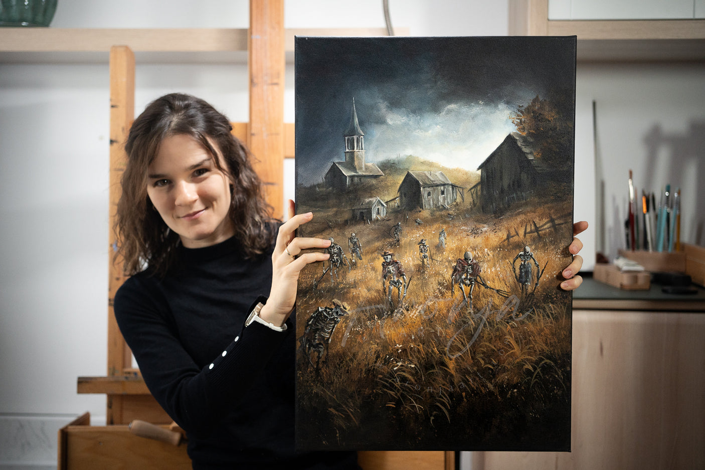 Woman artist holding SORCERY Gothic Original Painting "Raise Militia" - 60x40cm / Acrylics depicting a dark, fantasy landscape with militia figures in a field near rustic buildings, showcasing hand-painted traditional fine art inspired by gaming and nature. Fantasy Landscapes, Dragons, Creatures and gaming art.