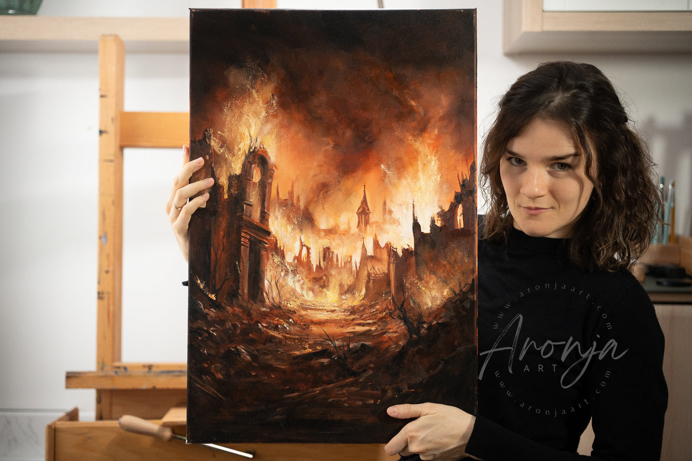 Woman holding SORCERY Gothic Original Painting "Raze" - 60x40cm / Oils, depicting a hand-painted fantasy landscape of fiery ruins, showcasing traditional oil painting skills by a fantasy artist.