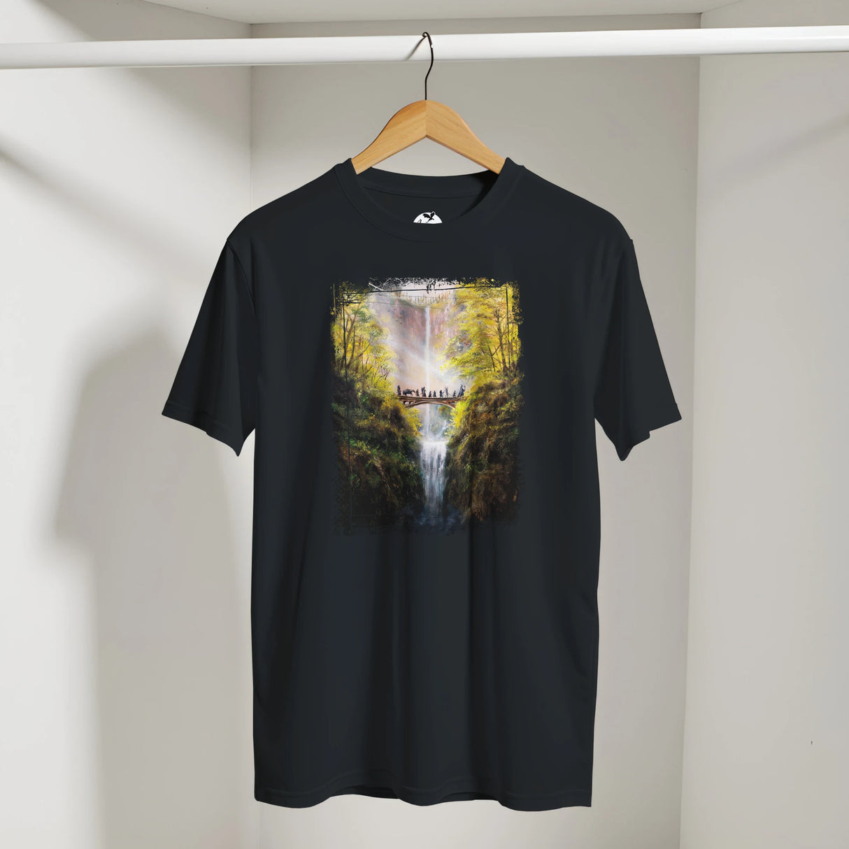 Black The Bridge T-shirt featuring a detailed fantasy art print of a wooden bridge over a waterfall surrounded by lush, mythical forest landscape in warm sunlight.